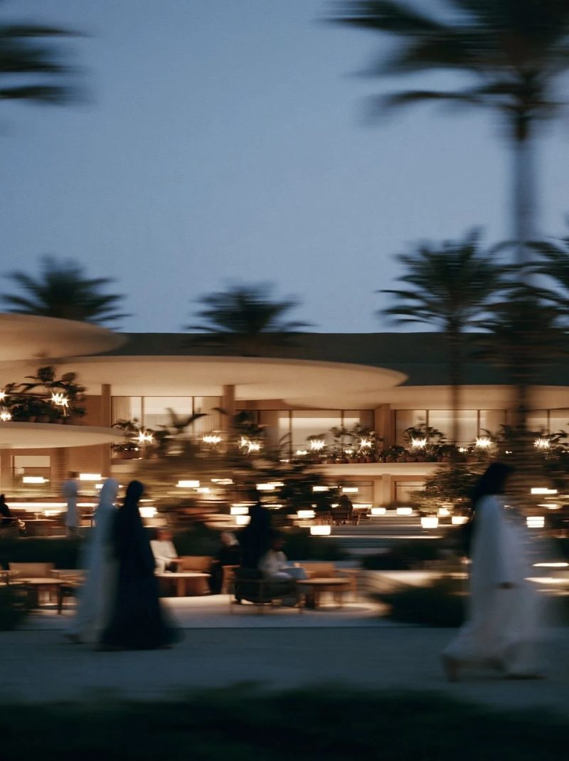 M11 · Árdh, concept image — a low curved pavilion lit at dusk among palms, figures walking past in motion blur