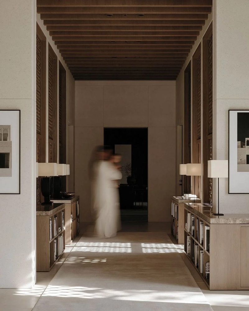 M18 · Árdh, concept image — a long timber-ceilinged corridor lined with low shelves and lamps, sunlight patterned on the floor, a figure in white walking through