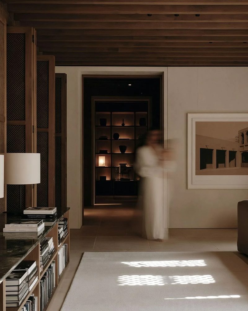M20 · Árdh, concept image — a view through a timber-framed room to a library beyond, low shelves and lamps, patterned sunlight on the floor, a figure in white